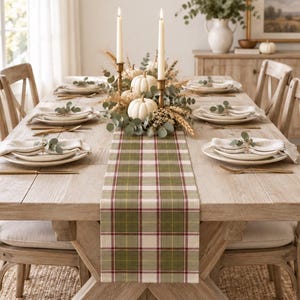 May include: A rectangular table runner featuring a plaid pattern in shades of olive green, cream, and thin burgundy lines. The runner is centered on a light-toned wooden dining table set with white plates, linen napkins, and gold-toned cutlery.