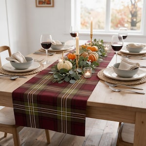 Puede incluir: Una mesa de comedor preparada con un camino de mesa de cuadros rojos, verdes y blancos. El centro de mesa incluye pequeñas calabazas blancas y naranjas, ramas de eucalipto y velas largas en candelabros dorados con portavelas de cristal.
