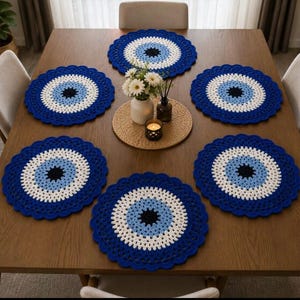May include: A set of six circular crochet placemats featuring a concentric eye pattern with a black center, light blue ring, white ring, and a dark blue scalloped outer edge. The placemats are arranged on a wooden table surface.