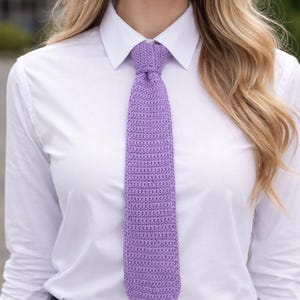 Könnte beinhalten: Eine lavendelfarbene Krawatte aus Häkelarbeit ist an einem weißen Hemd mit Kragen geknotet. Die Krawatte ist einfarbig und hat ein strukturiertes Aussehen. Das Outfit wird durch eine schwarze Hose ergänzt. Das Bild ist eine Nahaufnahme des Oberkörpers.