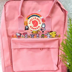 May include: A pink backpack with a front pocket and top handle. The backpack features an embroidered design of cartoon characters and the Fjallraven Kanken logo. The bag has a zipper closure and adjustable straps.