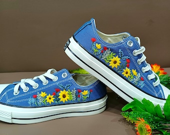 Puede incluir: Un par de zapatillas de lona azules con diseños florales bordados en amarillo, rojo y verde. Las flores están dispuestas en una línea a lo largo del lado de cada zapato.