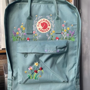 Könnte beinhalten: Ein teal-farbener Rucksack mit gestickten Blumenmustern, darunter Gänseblümchen, Lavendel und Pilze. Der Rucksack hat das Logo "Fjallraven Kanken" und den Namen "Elexia" auf der Vordertasche gestickt. Er hat einen oberen Griff und Schultergurte.