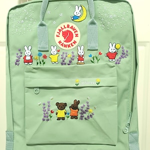 May include: A light green backpack with embroidered bunny and bear designs. The backpack features a front pocket, top handle, and adjustable straps. The embroidered designs include bunnies with flowers, a bear, and the text "Johanna".