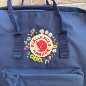 May include: Navy blue backpack with a front pocket and top handles. The backpack features a circular patch with the text "Fjallraven Kanken" and a red fox logo, surrounded by a floral and bee embroidery design.