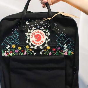 May include: Black backpack with embroidered mountain and floral design. The backpack features a round logo with a red emblem and white text. The embroidery includes a crescent moon, stars, and colorful flowers.