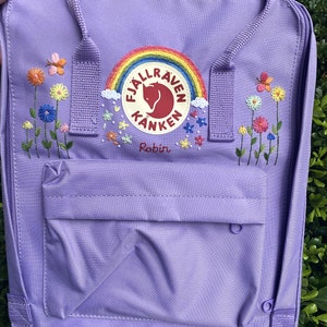 May include: Purple Fjällräven Kånken backpack with a rainbow and fox logo embroidered on the front. The backpack has a small pocket on the front and two adjustable straps. The backpack is decorated with embroidered flowers and the name "Robin" is written below the logo.
