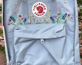 Puede incluir: Una mochila Fjällräven Kånken azul claro con un bolsillo frontal y flores y abejas bordadas. El logotipo es un zorro rojo dentro de un círculo con el texto "Fjällräven Kånken".