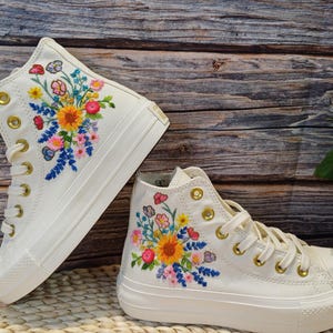 May include: A pair of white high-top canvas shoes with colourful embroidered flowers. The shoes have gold eyelets and laces.