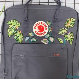May include: A gray Fjällräven Kånken backpack with a red fox logo and embroidered green and white tropical leaves. The backpack has a small pocket on the front with the word "POE" embroidered in red.