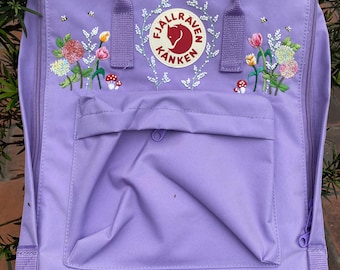Puede incluir: Una mochila Fjällräven Kånken de color morado con un diseño floral bordado en blanco y el logotipo de Fjällräven. La mochila tiene dos correas ajustables y un bolsillo frontal con cremallera.