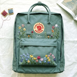 Könnte beinhalten: Ein tealfarbener Rucksack mit gestickten bunten Blumen. Der Rucksack hat eine Vordertasche mit Reißverschluss und einen runden Logoaufnäher. Der Name "Julie" ist unter dem Logo gestickt. Zwei Garnrollen befinden sich in der oberen linken Ecke.