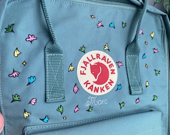 May include: A teal blue Fjällräven Kånken backpack with a red fox logo and the text "Fjällräven Kånken" embroidered on the front. The backpack is decorated with colorful hand-painted flowers and the name "Marc" is written in cursive on the logo.