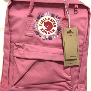 May include: A pink Fjällräven Kånken backpack with a floral embroidered logo. The logo features a fox and the words "Fjällräven Kånken".
