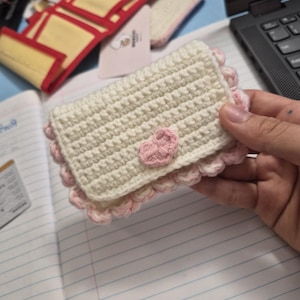 May include: A handmade, off-white crochet wallet with a pink heart accent. The wallet has a scalloped pink trim and a textured stitch pattern. The wallet is held in a hand, with a notebook and other items in the background.