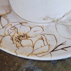 K&ouml;nnte beinhalten: Ein wei&szlig;er Filzhut mit einem braunen Blumenmuster, einschlie&szlig;lich einer gro&szlig;en Blume und &Auml;sten, die in den Stoff gebrannt sind. Der Hut hat eine braune Schnur, die um die Krone gebunden ist.