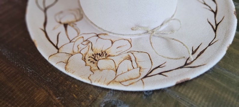 K&ouml;nnte beinhalten: Ein wei&szlig;er Filzhut mit einem braunen Blumenmuster, einschlie&szlig;lich einer gro&szlig;en Blume und &Auml;sten, die in den Stoff gebrannt sind. Der Hut hat eine braune Schnur, die um die Krone gebunden ist.