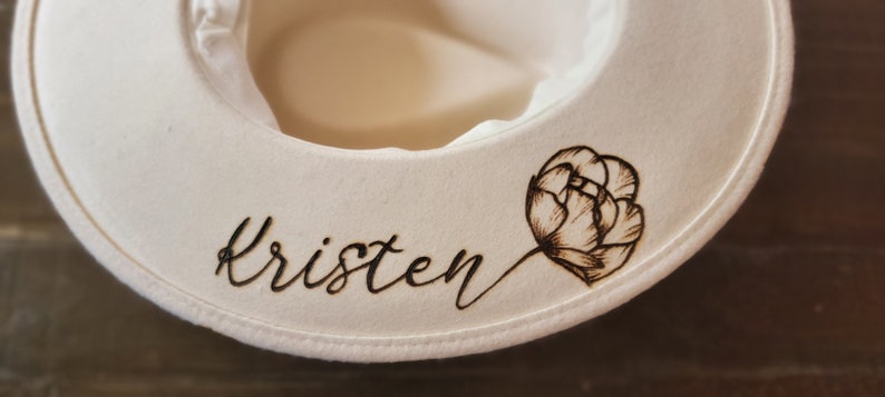 K&ouml;nnte beinhalten: Wei&szlig;er Sonnenhut mit dem Namen "Kristen" und einem schwarzen floralen Design, das in den Stoff gebrannt ist.