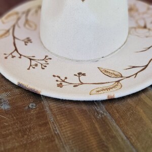 K&ouml;nnte beinhalten: Ein wei&szlig;er Filzhut im Fedora-Stil mit braunem Blumenmuster. Der Hut hat eine breite Krempe und eine leicht eingedellte Krone.