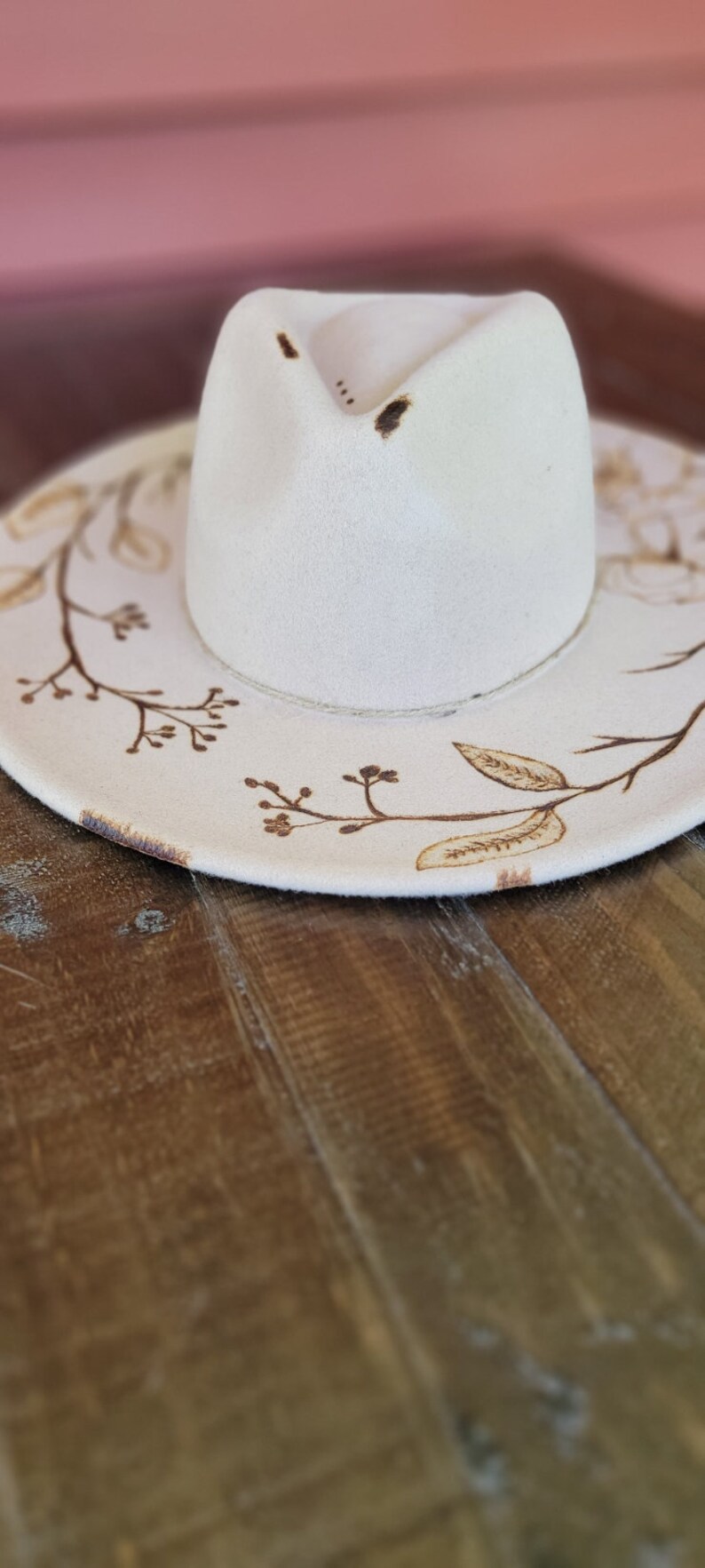 K&ouml;nnte beinhalten: Ein wei&szlig;er Filzhut im Fedora-Stil mit braunem Blumenmuster. Der Hut hat eine breite Krempe und eine leicht eingedellte Krone.