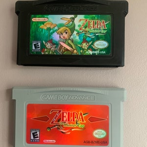 May include: Two Game Boy Advance video game cartridges for The Legend of Zelda: The Minish Cap. The top cartridge is black with a green and yellow label. The bottom cartridge is grey with a red and yellow label. Both cartridges have the game title and the PEGI rating of "3".
