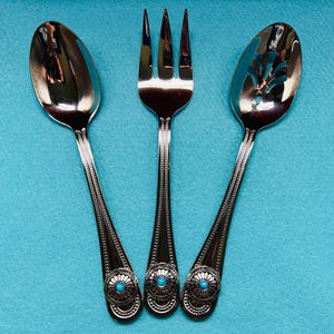 May include: A set of three silver serving utensils: a spoon, a fork, and a slotted spoon. Each handle has a decorative design with a turquoise-colored center. The utensils are arranged on a turquoise background.