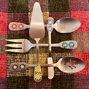 May include: A collection of stainless steel serving utensils with decorative floral accents. Includes a cake server, fork, slotted spoon, and spoons. The handles feature red, blue, and yellow flower designs. The utensils are arranged on a colorful, patterned surface.
