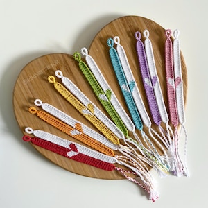 May include: A collection of colourful friendship bracelets with white hearts woven into the design. The bracelets are arranged in a fan shape on a wooden heart-shaped surface.