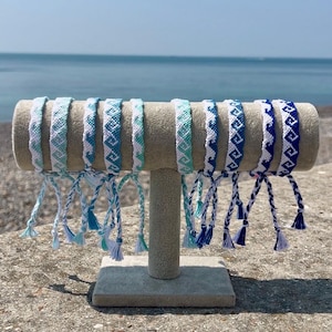 May include: A collection of eight woven friendship bracelets displayed on a beige T-bar stand. Each bracelet features a repeating geometric wave pattern in varying shades of blue, teal, and white, finished with braided ties and small tassels.