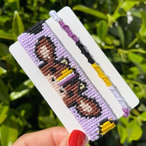 May include: Two friendship bracelets, one with a purple, yellow and black pattern and the other with a detailed woven design featuring brown, black, yellow and white threads on a lavender background.