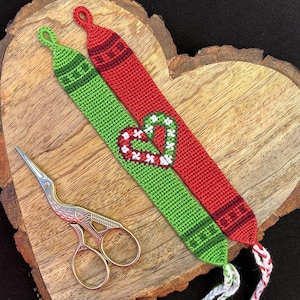 May include: Two friendship bracelets, one green and one red, with a white heart design. The bracelets are tied together and are on a wooden heart-shaped surface. A pair of gold scissors are also in the image.