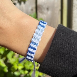 May include: A woven friendship bracelet featuring a repeating pattern of dark blue, light blue, and white threads. The bracelet has a textured, knotted design with a consistent width and a secure, flat construction that wraps around the wrist.