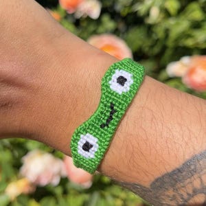 May include: A woven friendship bracelet made of green thread featuring a frog face design. The bracelet includes two white circular eye patches with black pupils and a small black stitched mouth centered between the eyes on a green band.