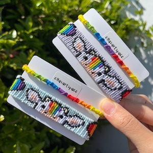 May include: Two woven friendship bracelets on white display cards. Each features a pixelated cat design with rainbow accents and a thin, multi-coloured braided cord. The text "ellasartspace" is printed on each card. The bracelets use vibrant thread.
