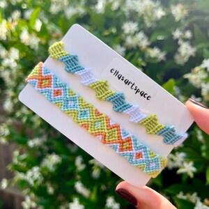 May include: Two woven friendship bracelets attached to a white display card with the text "ellasartspace". The top bracelet features a zigzag pattern in lime green, teal, and white. The bottom bracelet displays a diamond pattern in orange, light blue, lime green, and white.