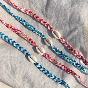May include: Three woven friendship bracelets featuring chevron patterns in vibrant colours including red, orange, yellow, teal, and blue. Each bracelet is accented with a single white cowrie shell centred along the braided cord construction.