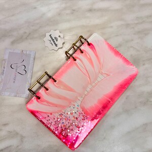 May include: A handmade, pink and white resin notebook with gold-colored binding. The notebook features a floral design and glitter accents. A tag that says "handmade for you" is attached.