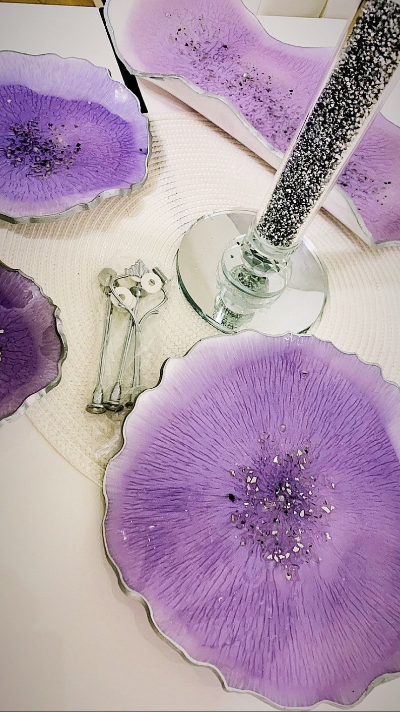 K&ouml;nnte beinhalten: Lila Resin-Untersetzer mit silbernen R&auml;ndern und eingebetteten Glitzerpartikeln. Das Set enth&auml;lt runde und unregelm&auml;&szlig;ig geformte Untersetzer, einen dekorativen Glasstiel und Servierutensilien. Die Untersetzer sind helllila.