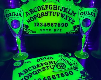 Könnte beinhalten: Ein neongrünes Ouija-Brett-Set mit zwei passenden Sektgläsern. Das Brett zeigt das Alphabet, Zahlen und die Wörter "Ja", "Nein" und "Auf Wiedersehen". Die Gläser und das Brett leuchten unter Schwarzlicht.