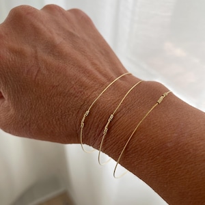 Può includere: Tre braccialetti sottili in filo d'oro, ognuno caratterizzato da un piccolo dettaglio in filo avvolto e testurizzato lungo la fascia. I braccialetti sono di forma circolare e hanno una finitura metallica lucida, indossati insieme al polso.