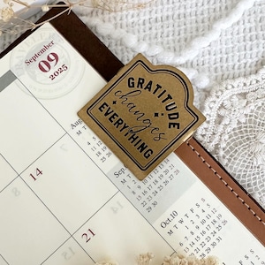May include: A gold-colored sticker with the text "Gratitude changes everything" on a brown leather planner with a calendar page for September 2025.