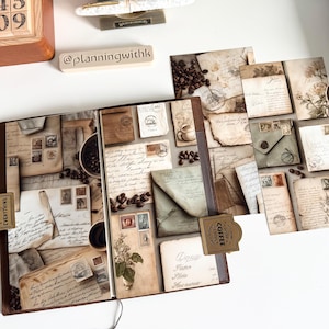 May include: An open journal with vintage-style pages featuring coffee beans, stamps, and handwritten letters. The journal is brown with a leather-like texture. The image also shows pens, a wooden stamp holder, and a small plant in a white pot.