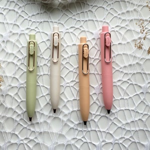 May include: Four pastel coloured pens with metal clips. The pens are green, white, orange, and pink. The pens are arranged in a row on a white crocheted surface.