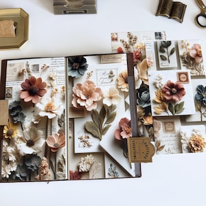 May include: An open journal with paper flowers and decorative elements. The journal features a variety of paper flowers in shades of white, peach, and brown. A small tag reads "Gratitude changes everything."