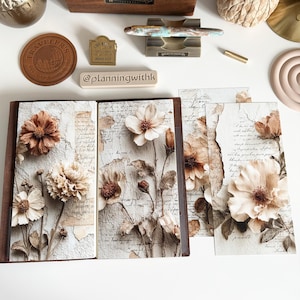 May include: A collection of stationery and desk accessories, including a wooden pen holder, decorative objects, and a journal with floral designs. The journal features cream-colored pages with dried flowers and script. The color palette is neutral with brown and cream tones.