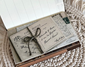 May include: A vintage-style journal with a brown leather cover and a stack of old postcards on top. The postcards have handwritten messages and stamps. The journal is open to a blank page with a calendar for January 2024.