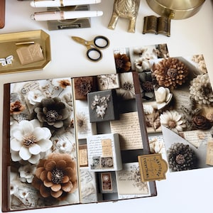 May include: An open scrapbook with floral and vintage ephemera designs. The pages feature large paper flowers in shades of brown and white, with a decorative tag that reads "Gratitude changes everything". Other items include pens, scissors, and a small gold deer figurine.