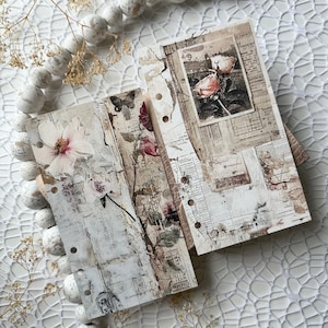 May include: Three white pages with a distressed, vintage look. Each page features a different floral design, including a pink rose, a white flower, and a butterfly. The pages are bound together with metal rings.