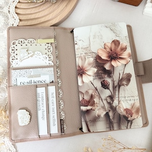 May include: Open planner with a light brown leather cover, featuring a floral print insert and multiple card slots. The planner includes cards with text like "re-sil-ience" and "Self-care isn't selfish". A decorative coffee cup charm is visible.