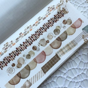 May include: A journal page with several decorative washi tape designs. The tapes feature patterns like brick, circles, and plaid in shades of brown, beige, and white. The page is open, and the tapes are arranged across the spread.